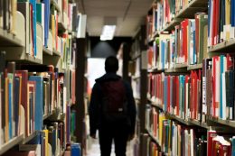 Student with a backpack walking away from library books shelves into darkness.