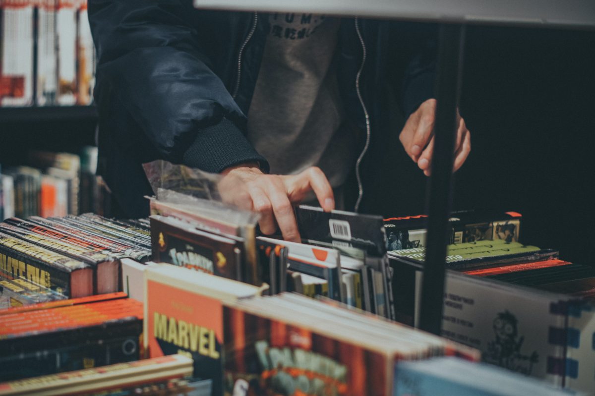 Man in a bomber jacket and grey t-shirt is reaching into a low bin of comics. We see he is in a used book store surrounded by comics and graphic novels.